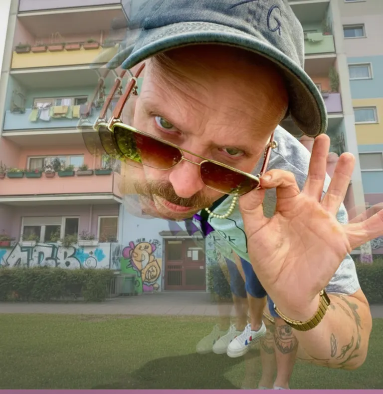 Roger G. lehnt sich mit Cap und Sonnenbrille im Fisheye-Look vor einem pastellfarbenen Plattenbau ins Bild.